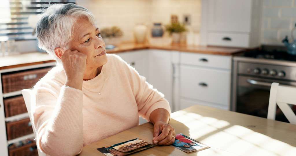 Older adult seated at a kitchen table, looking off to the side while holding and viewing printed photographs, with sunlight coming through a nearby window