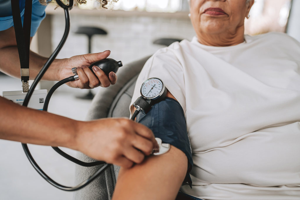 Close-up of a person seated indoors while another person checks blood pressure using a cuff wrapped around the upper arm and a manual pump with gauge