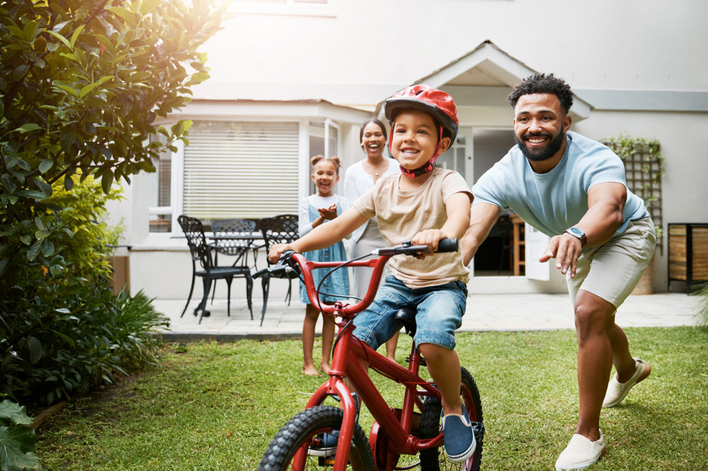 Child wearing a helmet rides a bicycle in a backyard while an adult runs alongside, demonstrating safe biking practices